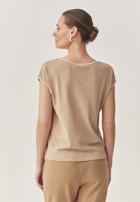 Top beige a maniche corte con scollo a V e profilo bianco. Texture liscia. Indossato con pantaloni beige abbinati. La vista posteriore mostra la forma e la vestibilità delle spalle.