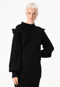 Black ribbed knit sweater with a high neck, ruffle accents on shoulders, and long balloon sleeves, featuring a fitted silhouette.