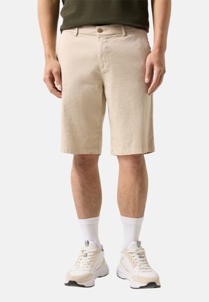 Mężczyzna w beżowych szortach do kolan, białych skarpetach i białych trampkach, stojący na jednolitym jasnym tle.