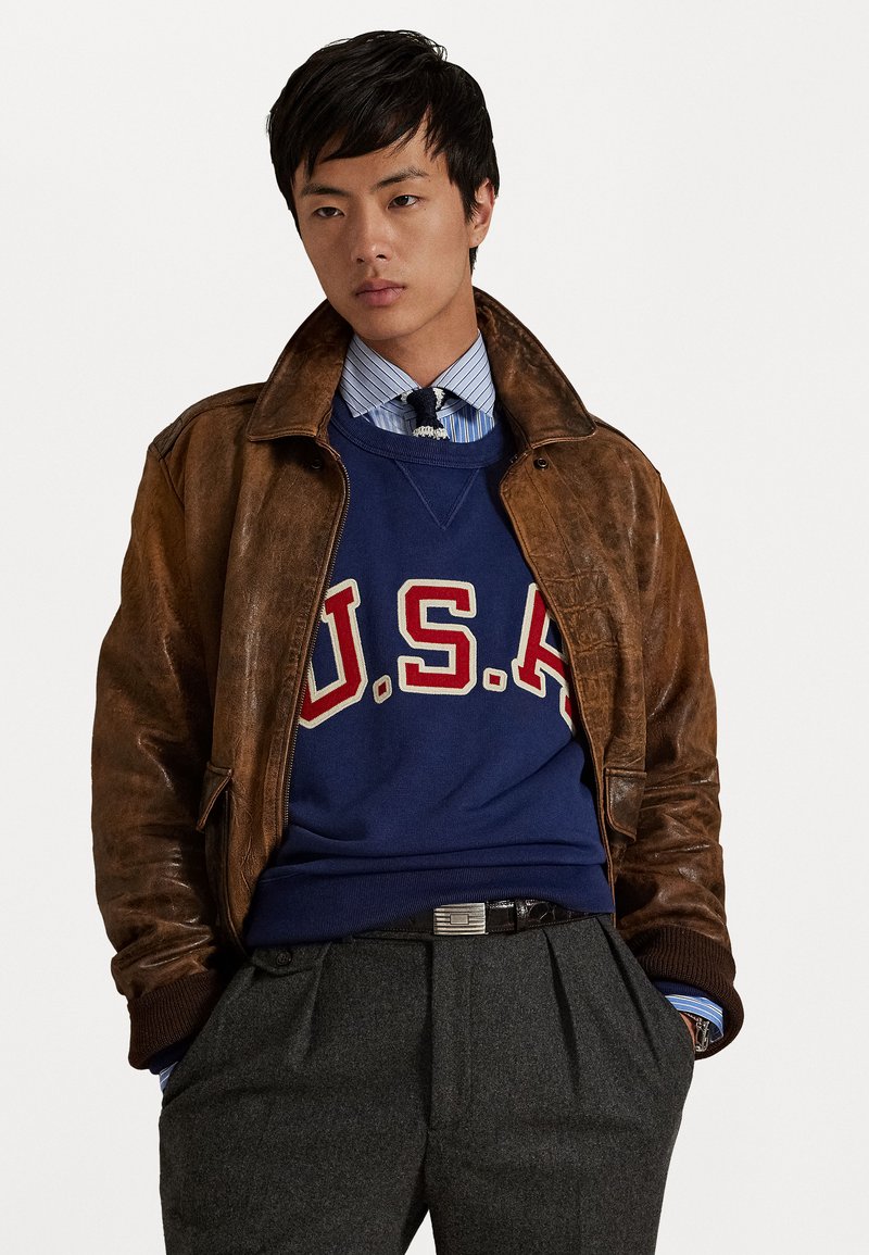Veste en cuir marron portée par-dessus un sweat à capuche bleu avec "U.S.A." en rouge, associé à une chemise rayée. Un pantalon gris plissé termine la tenue.