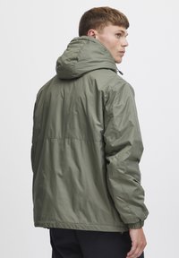 Oliven grønn lett jakke med hette, elastiske mansjetter og en litt oversized passform. Glatt stoff med vanntett finish.