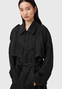 Svart trenchcoat med kort design, stor krage, dubbelradiga knappar, avtagbart midjebälte och sidofickor. Slät textur.