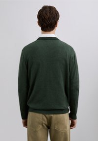 Suéter de punto verde oscuro con diseño sencillo, mangas largas y puños acanalados, combinado con pantalones beige claro.