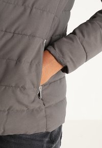 Grå dunjakke med quiltet tekstur, der har en lynlåslomme og en tætsiddende manchet. Materialet virker syntetisk og glat.