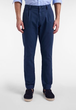 Marineblauwe katoenen broek met een rechte pijp, voorzien van voorplooien en een gladde textuur. Draagt donkerkleurige instapschoenen.