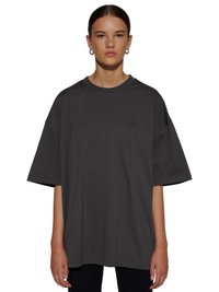 Jeune femme aux cheveux foncés portant des boucles d’oreilles anneaux argentées et un T-shirt noir oversize avec un petit logo brodé sur la poitrine.