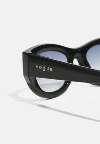 Črne plastične sončne očala z debelim okvirjem, gradientnimi modrimi lečami in imenom blagovne znamke "vogue" vgraviranim na strani.
