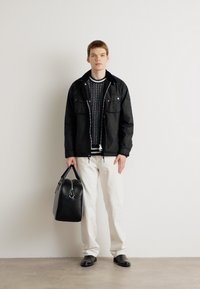 Black zip-up jacket with two chest pockets, layered over a striped sweater, paired with white trousers and black shoes, holding a black bag.