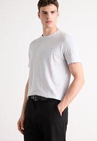 T-shirt léger gris clair à col rond en tissu doux, avec une texture subtile et un petit logo sur la poitrine, associé à un pantalon noir et une ceinture.