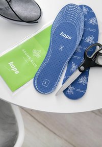 Modré perforované stélky s květinovými vzory, položené vedle nůžek a zeleného obalu, na kulatém bílém stole.