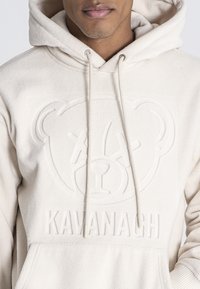 Kremowa polarowa bluza z kapturem z wytłoczonym logo niedźwiedzia i napisem "Kavanagh" z przodu, z regulowanym sznurkiem i kieszenią kangurką.