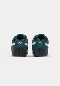 Puma tossud, tumeroheline sametülem koos valgete triipudega ja mustade kummitaldadega. Kollased logo aktsendid kannal täiendavad disaini.