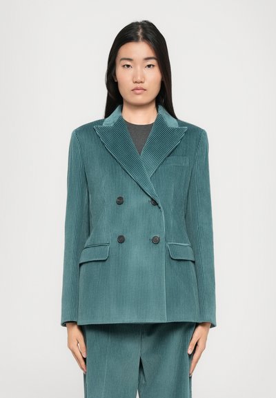 Mujer vestida con un blazer de pana de color teal de doble botonadura y pantalones a juego, de pie contra un fondo claro liso, mirando al frente con una expresión neutral.