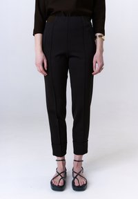 Zwarte op maat gemaakte pantalons met plooien, enkellange boorden en een gladde textuur, gecombineerd met zwarte sandalen met bandjes. Eenvoudig en modern ontwerp.