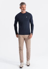 T-paita tummansinisestä kankaasta, jossa on pyöreä kaula-aukko, yhdistettynä beigeihin housuihin ja ruskeilla yksityiskohdilla varustettuihin tennareihin. Teksturoitu kangas, minimaalinen logo.