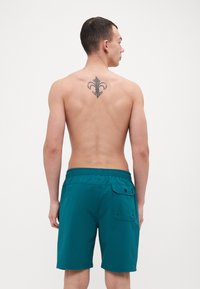 Mėlynos maudymosi šortai su elastine juosta, šoniniu kišenėmis ir matomomis siūlėmis. Modelis parodo nugaros vaizdą, atskleidžiančią tatuiruotę ant viršutinės nugaros.