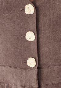 Bruine stof met een gestructureerde weving bevat drie schelpvormige beige knopen met dunne lijnen, die detail toevoegen aan het ontwerp en de sluiting.