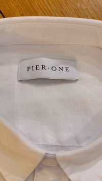 Tessuto di camicia bianca con una texture morbida, dotato di colletto e di un'etichetta etichettata "PIER ONE" in nero su bianco. Bottone visibile sotto il colletto.