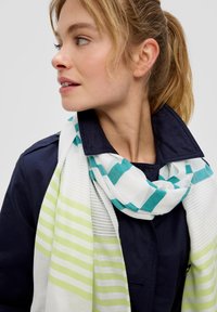 Écharpe légère blanche avec des rayures vertes et bleu-vert, présentant un motif à carreaux. Portée sur une veste bleu marine foncée. Texture douce.