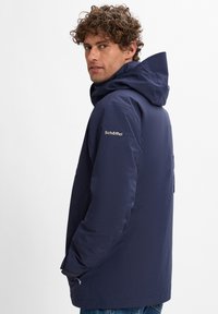 Navy wasserdichte Jacke mit Kapuze, Seitentaschen und dem "Schöffel"-Logo am Oberarm. Glatte Textur und gerade Schnittform.