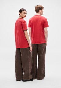 Camisetas de algodón rojas con mangas cortas, combinadas con pantalones marrones de pierna ancha. Texturas suaves y diseño minimalista, visibles desde la vista trasera.