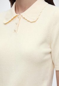Haut à manches courtes couleur crème avec col festonné et trois petits boutons en perle sur la patte de boutonnage devant.