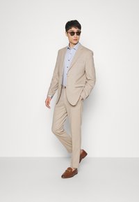 Béžový oblek so slim fit strihom, s klopami, svetlo modrá košeľa s malým karovaním a hnedé mokasíny. Doplnky zahŕňajú tmavé slnečné okuliare.