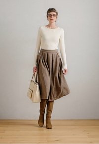 Barna plisszírozott szoknya, krémszínű bordás hosszú ujjú felső, magas barna csizma és egy krémszínű kézitáska. A modell egy világosszürke háttér előtt áll.