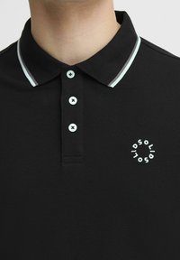 Svart poloskjorte laget av teksturert stoff. Har en krage med lyseblå detaljer og tre hvite knapper. Har en liten sirkulær logo på brystet.