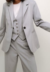 Costume gris clair comprenant un blazer avec revers en pointe, un gilet boutonné et un pantalon à jambes larges. Texture lisse, coupe ajustée, design minimaliste.
