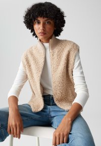 Gilet in pile beige con colletto a costine, dotato di chiusura con zip e maniche corte. Indossato sopra una maglietta a maniche lunghe bianca e jeans blu.