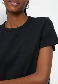 Gros plan d'une personne souriante portant un t-shirt noir uni à manches courtes et un col rond, ainsi que des boucles d'oreilles en anneau, sur un fond clair.