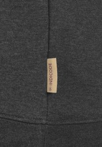 Tela de sudadera con capucha en gris oscuro con un acabado texturizado. Una etiqueta color canela en el lateral muestra "INDICODE" en letras burdeos.