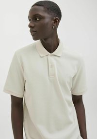 Polo de color beige claro con tejido texturizado, de mangas cortas y cuello clásico; cuenta con tres botones y un discreto detalle de logo.