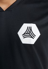 Camiseta atlética negra con cuello en V, que presenta un prominente logo hexagonal blanco. La tela parece suave con un ligero brillo.