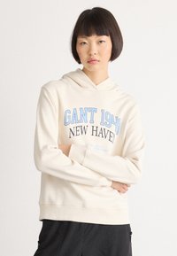 Person with short dark hair wears cream hoodie with "GANT 1949 NEW HAVEN" text, paired with black pants, standing against white background.