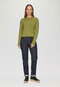 Grüner Langarmpullover kombiniert mit dunklen Jeans und schwarzen Sneakers. Die Jeans verfügen über gestickte Details und umgeschlagene Säume.