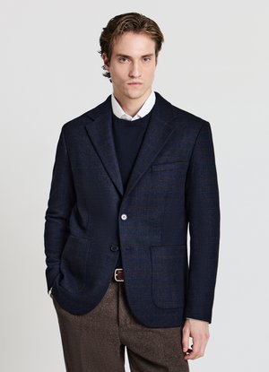 Marineblazer mit strukturiertem Muster, zwei aufgesetzten Taschen und einem Ein-Knopf-Verschluss, über einem dunklen Pullover und einem weißen Hemd getragen.