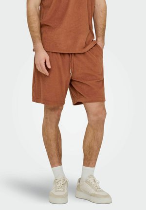 Mand iført rustfarvede frottéshorts og matchende top, stående med den ene hånd i lommen, hvide sokker og beige sneakers.