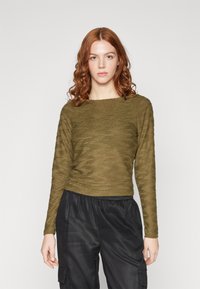 Top lung cu mânecă lungă, texturat, de culoare verde măsliniu, cu un decolteu larg, asociat cu pantaloni cargo negri, cu talie elastică, prezentând o croială relaxată.