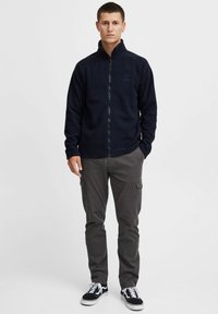 Marine fleece zip-up jacket met hoge kraag en lange mouwen. Draagt men met grijze cargobroek en zwarte sneakers met witte veters. Eenvoudig ontwerp.