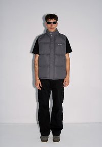 Szara kamizelka puchowa z wysokim kołnierzem, czarna koszulka z krótkim rękawem, czarne spodnie cargo, srebrne okulary przeciwsłoneczne i szare obuwie na jednolitym tle.