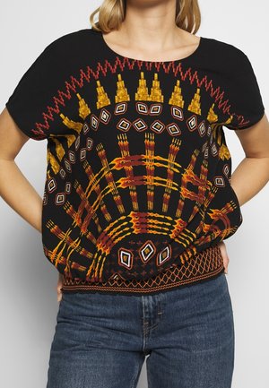 Personne portant un haut noir à manches courtes avec des motifs géométriques vifs orange, rouge et jaune et un jean bleu, les mains posées sur les hanches.