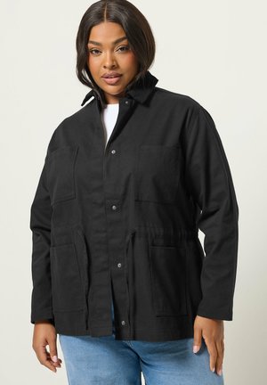 Nainen, joka käyttää mustaa napillista takkia suurilla taskuilla ja kiristysnarulla, yhdistettynä vaaleansinisiiin farkkuihin, seisoo yksinkertaisen taustan edessä.