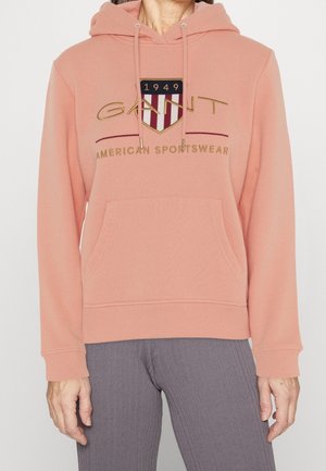 Personne portant un sweat à capuche GANT rose saumon avec poche kangourou et pantalon côtelé gris, cadrée à mi-torse sans montrer le visage.