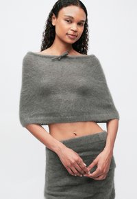 Ženska z kodrastimi lasmi, oblečena v puhast sivi crop top z rameni in ujemajočo se krilo z visokim pasom, stoji pred enostavno svetlo ozadje.