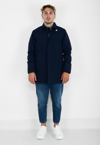 Giacca impermeabile blu navy con chiusura a zip, maniche lunghe e tasche laterali. Indossata con jeans in denim azzurro e sneaker bianche.