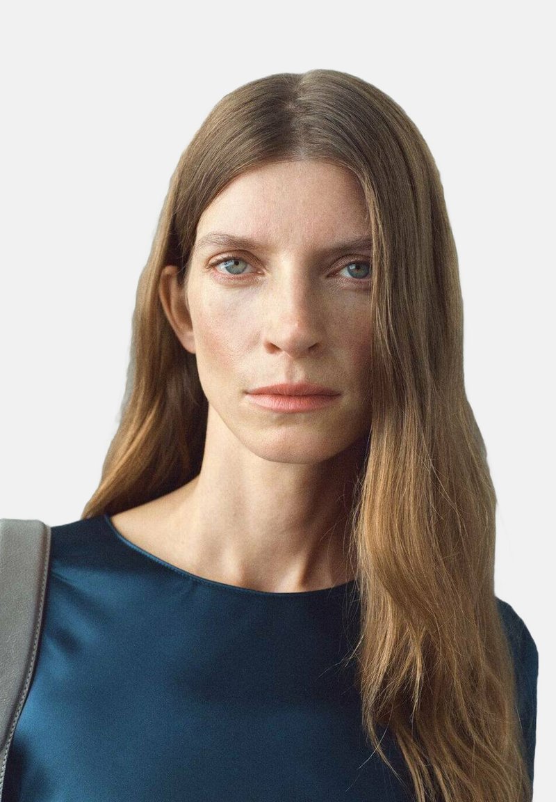 Vrouw met lang lichtbruin haar, blauwe ogen, die een donkerblauwe top draagt en een grijze schoudertas vasthoudt, tegen een effen lichte achtergrond.