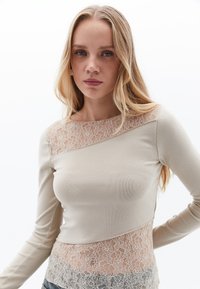 Beige Langarmtop mit strukturiertem Spitzenbesatz auf der oberen Brust und am unteren Saum, mit einer taillierten Passform und glatter Stoffoberfläche.