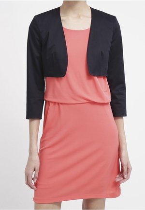 Robe corail associée à un blazer noir court. La robe a une coupe ample, tandis que le blazer possède des manches trois-quarts et une texture lisse.